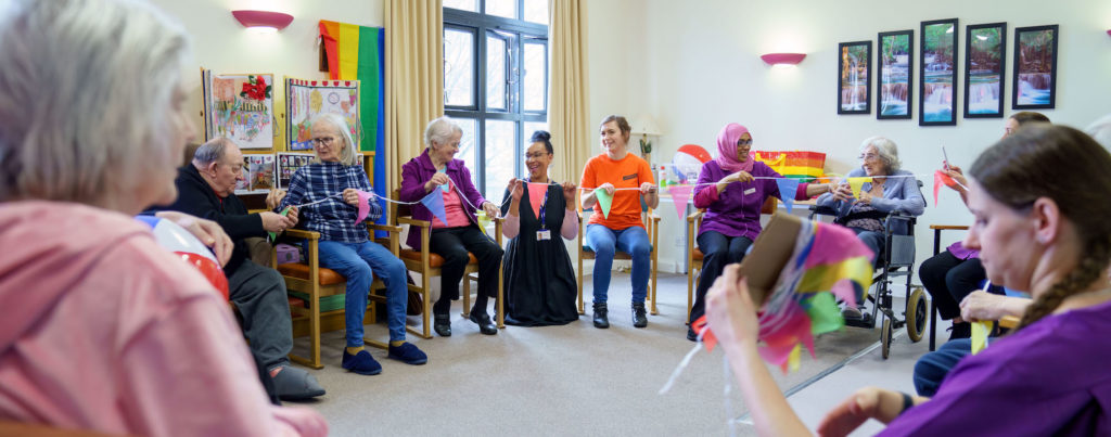 A group of older people and care home staff sit in a circle and run bunting between them, smiling