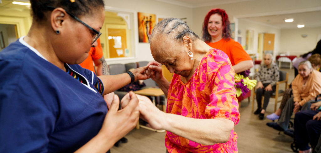 A care home staff member helps an older person to walk across the room by holding her hands